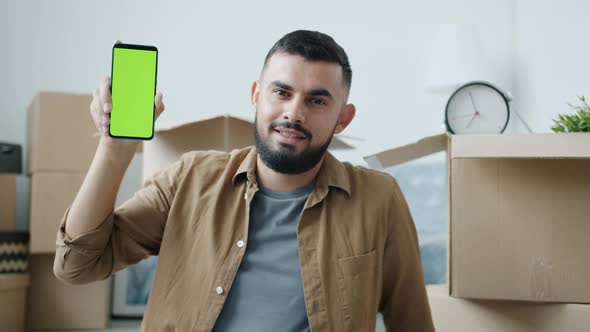 Portrait of Joyful Arab Guy Holding Green Screen Smartphone in New Apartment with Boxes alt