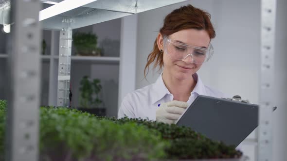 Smiling Female Biology Laboratory Worker Examines the State of Micro Greens in Containers in Modern alt