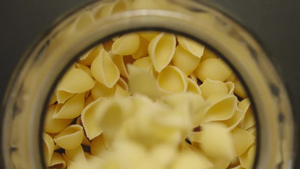 TOP VIEW: Uncooked Conchiglie Pasta Fall Into Glass Jar On Black Background - Slow Motion alt