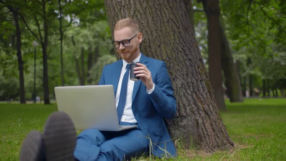Young Businessman with Laptop Sitting Under Tree Drinking Coffee and Working Remotely in Park alt
