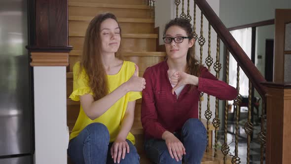 Skeptic Young Woman in Eyeglasses Showing Thumb Down As Cheerful Twin Sister Gesturing Thumb Up alt