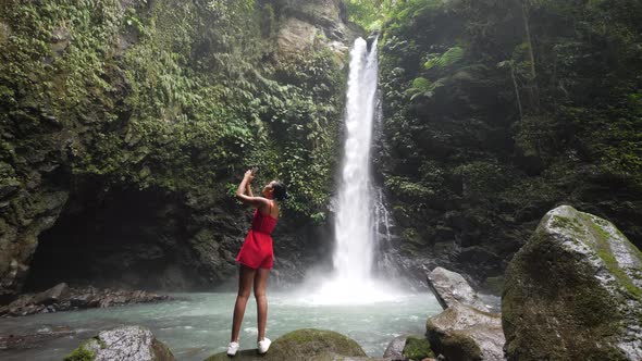 A Young Tourist Standing at a Waterfall Capturing Photos Using a Smartphone alt