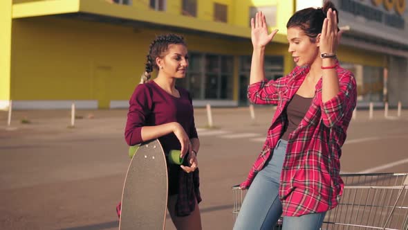 Two Hipster Girls Standing By the Shopping Cart on Parking By the Citymall During Sunset alt
