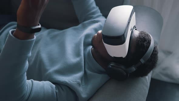 AfricanAmerican Man Wearing Virtual Reality Glasses and Headset Lying on the Sofa alt