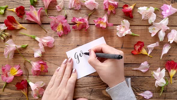 Writing GOOD MORNING on a card near flowers top view alt