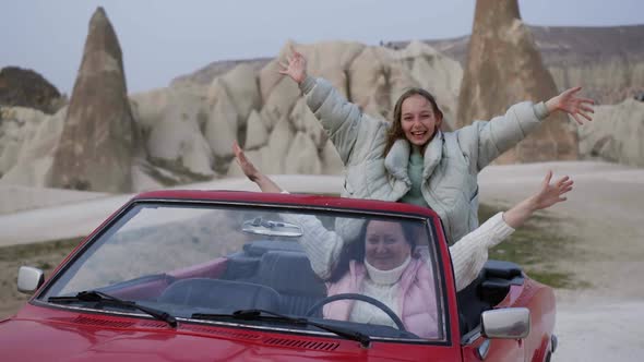 Happy Woman and Playful Girl Swaying Hands in Red Retro Cabrio Car Against Rock Formations