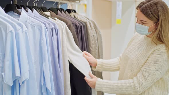 a Young Woman in a Protective Mask in a Clothing Store Chooses Casual Clothes Hung on Hangers alt