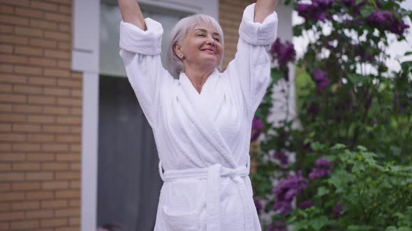 Excited Mature Woman Stretching in the Morning Standing on Backyard Porch Smiling Gesturing Thumbs alt