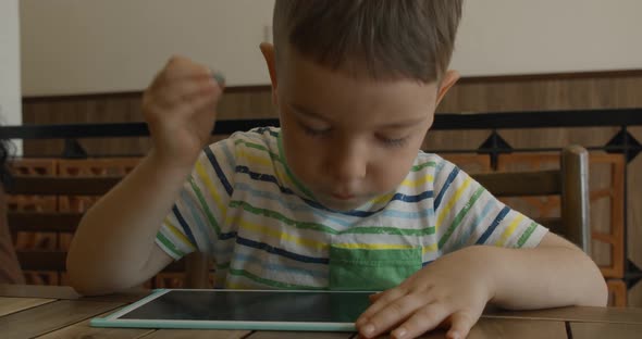 A Portrait of a Child Sits at a Table at Home and Draws on His Tablet with a Special Pen a alt
