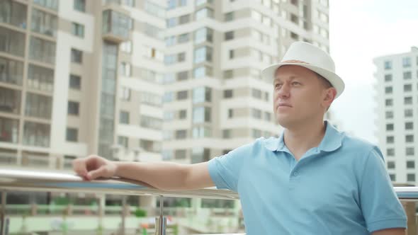 A Portrait of a Man in a Hat Stands By a Metal Fence Put His Hand on the Railing Looks Around alt