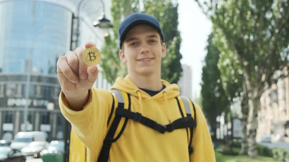Handsome Young Guy Shows a Golden Bitcoin in His Hand and Smiles alt