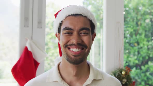 Portrait of happy biracial young man in santa hat at home during christmas alt