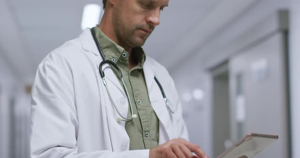Thougtful caucasian male doctor using digital tablet in the corridor at hospital alt