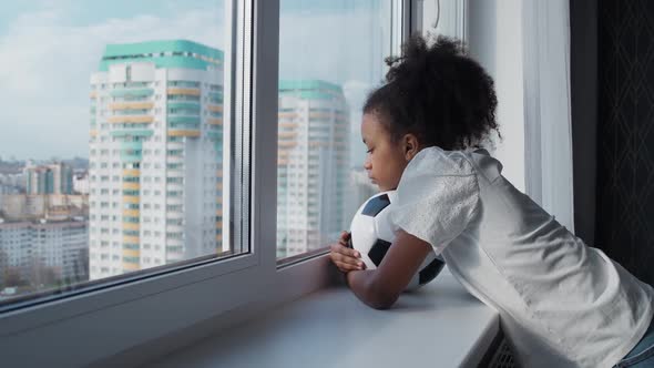 Handheld African Girl on a Home Quarantine Bored Girl Looking Out the Window and Holds a Soccer Ball alt