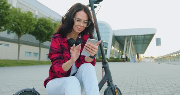 Woman with Headphones which Reviewing Funny Videos on Mobile while Sitting on Electric Scooter alt