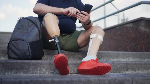 Unrecognizable Man with Prosthetic Legs Sitting on Urban City Stairs Messaging Online on Smartphone alt