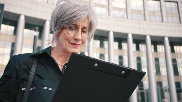 Attractive Woman with Flawless Makeup Looks at Documents in a Folder While Standing on a City Street alt