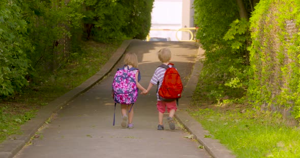 Two kindergarten students walking along a path to school together holding hands alt
