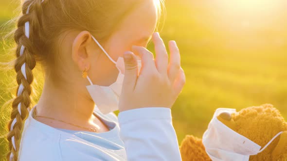 Crying Girl in Medical Protective Mask Holding a Teddy Bear at Sunset alt