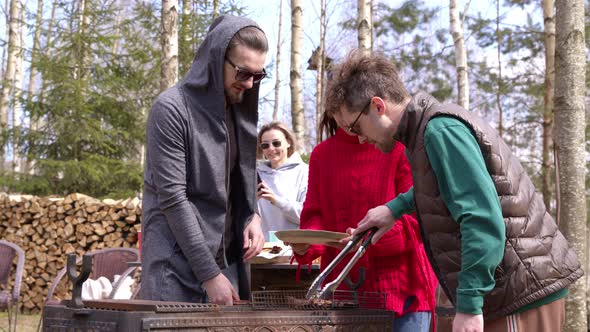 Young People Take Grilled Meat Off Grill, Put Steaks on Plates. Picnic Campfire