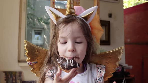 A Cute Little Girl at a Party in a Unicorn Costume Bites Off a Sweet Donut with Chocolate Cream alt