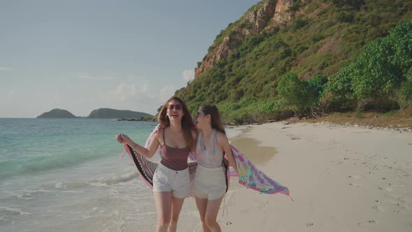 Two Asian women walking on the beach with a scarf in their hands.