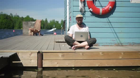 Young American Man is Working with Laptop While Sitting on Lake Pier on Summer Day Spbd alt