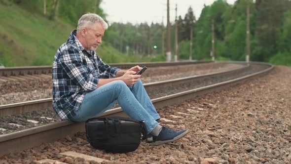 An Elderly Male Tourist Sitting on the Railway Tracks Looking at the Information on the Phone and