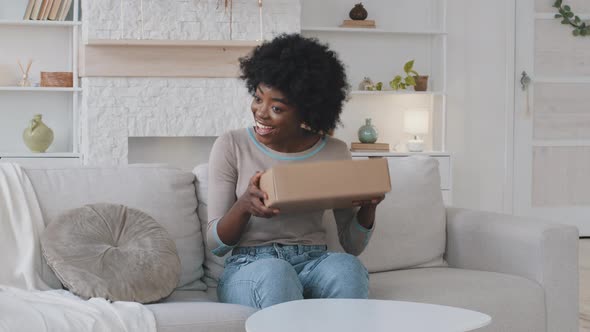 Smiling Happy African Young Woman Customer Sit on Sofa Shakes Parcel Carton Box Satisfied with alt
