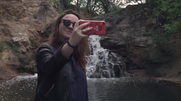Girl in Sunglasses Taking a Selfie Against the Backdrop of a Small Waterfall
