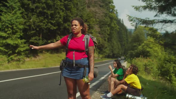 Tired African American Female Backpackers Hitchhiking on Highway During Mountain Hiking alt
