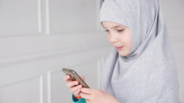 Portrait of Young Muslim Teen Girl in Grey Hijab Is Playing Smartphone alt