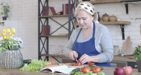 Portrait of Concentrated Senior Woman Cutting Tomato in Modern Kitchen. Positive Middle-aged alt