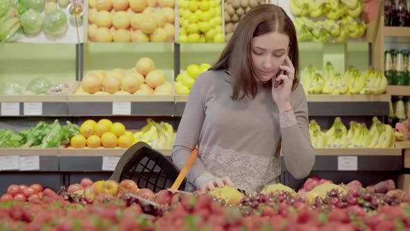 Portrait of Pretty Young Woman with Shopping Basket Choosing Fruits in Grocery and Talking on the alt