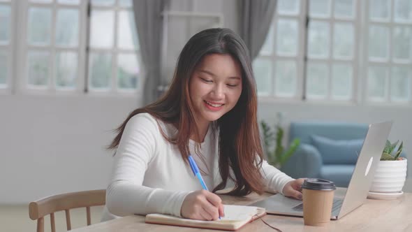 Happy Asian Girl Using Laptop Computer And Writing On Notebook At Home alt