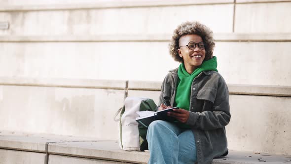 Laughing adult African woman wearing casual clothes writing something in notepad alt