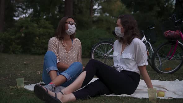 Two Young Women in Protective Face Masks Talking and Laughing While on a Picnic alt