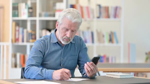 Old Businessman Using Smartphone and Doing Paperwork  alt