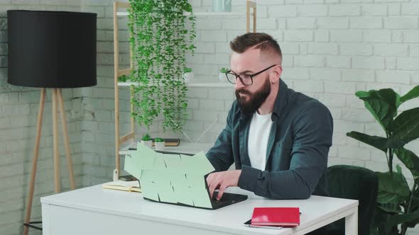 Bearded Man Types at the Laptop and Write Notice on the Paper Notebook alt