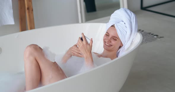 Young Charming Woman with a White Terry Towel on Head Lying Naked in Bathtub Smiles Using Mobile alt