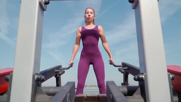 Young Athletic Woman in a Purple Sports Suit Doing a Squat Exercise on a Simulator in a City Park alt