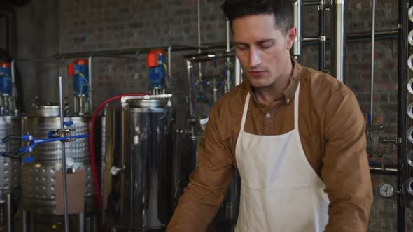 Portrait of serious caucasian man working at gin distillery, using equipment and looking to camera alt