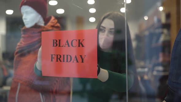 Young Brunette Woman in Covid-19 Face Mask Hanging Black Friday Sales Announcement on Shop Window alt