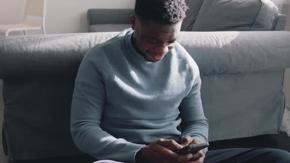 African American Black Man Sitting on the Floor in a Cozy Room at Home Using His Cell Phone alt