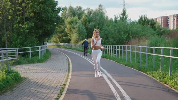 Beautiful Blonde Girl in a White Top and Leggings Jogging in the Park on a Sunny Day