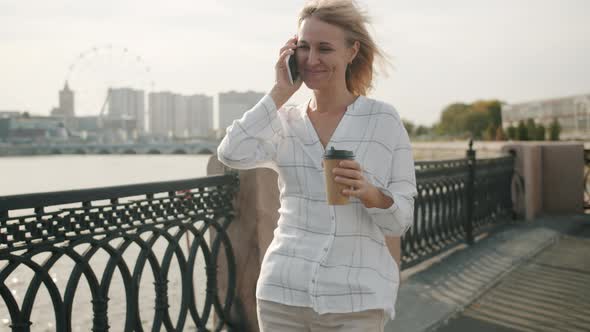 Cute Blonde Chatting on Mobile Phone Laughing Holding to Go Coffee Walking Outdoors in River alt