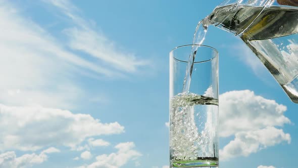 Pour Water Into A Transparent Glass Against The Background Of The Sky, Beautiful Fluffy Clouds.
