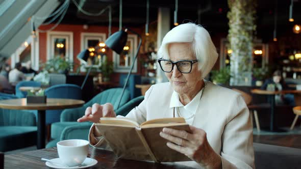 Smart Senior Business Lady Reading Book at Table in Restaurant Relaxing During Coffee Break alt