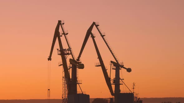 two construction cranes are working in the port at sunset