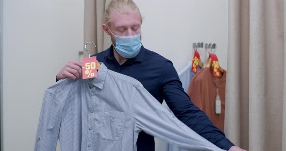 Middle Shot of Young Caucasian Male Shopper in Covid-19 Face Mask Trying on Shirt on Hanger alt